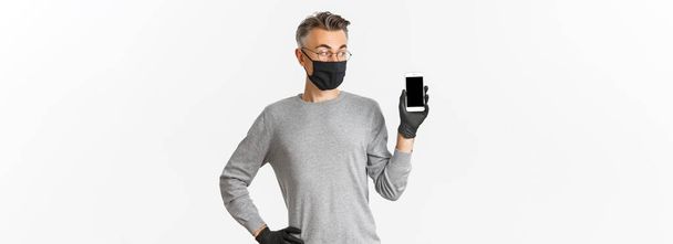 Concept of covid-19, social distancing and lifestyle. Portrait of handsome middle-aged man in medical mask, gloves and glasses, showing mobile phone screen, standing over white background. - Photo, Image