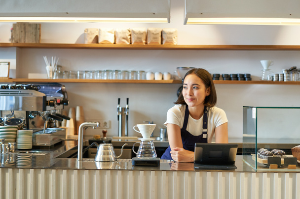 Szczęśliwy uśmiechnięty azjatycki barista, dziewczyna za ladą, praca z terminalem POS i zestaw filtrów warzących, parzenie kawy w kawiarni. - Zdjęcie, obraz