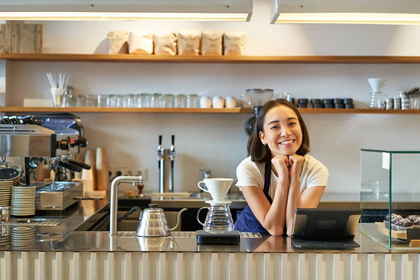 Café y pequeña empresa. Barista asiático sonriente en delantal, parado detrás del mostrador en la cafetería, trabajando con el terminal de punto de venta y el kit de preparación de filtro. - Foto, Imagen