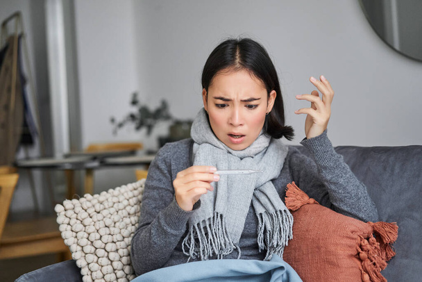 Mujer conmocionada mira preocupado por su termómetro, resfriado, tiene fiebre, sentado en licencia por enfermedad en casa. - Foto, Imagen