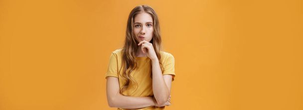 Сложный выбор. Портрет сосредоточенности серьезная творческая молодая женщина с волнистыми волосами ухмыляясь держа руку на подбородке в вдумчивой позе, принимая решение, думая над оранжевым фоном. - Фото, изображение