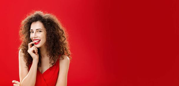 Μια φωτογραφία της σέξι και αισθησιακής ενήλικης κοπέλας του. με σγουρά μαλλιά σε κομψό φόρεμα δάγκωμα δάχτυλο και χαμογελώντας flirty αποπλάνηση καυτό άντρα κατά τη διάρκεια της επίσημης εκδήλωσης ποζάρουν κατά κόκκινο φόντο ρομαντικά. - Φωτογραφία, εικόνα