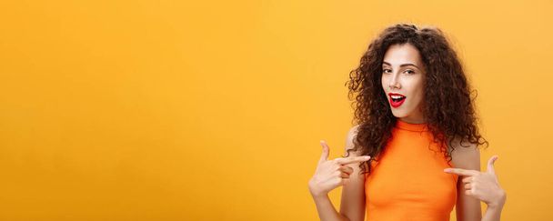 Proud and satisfied cool urban female with red lipstick. and curly hairstyle pointing at herself with self-assured expression winking bragging about skills and achievements over orange background. - Photo, Image