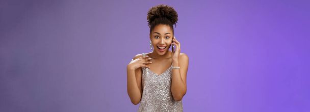 Εντυπωσιασμένη ομιλητική γοητευτική Αφροαμερικανή γυναίκα με γυαλιστερό ασημί βραδινό φόρεμα που δείχνει τον εαυτό της διασκεδασμένη έκπληκτη χαμογελώντας μιλώντας smartphone διευρύνει τα μάτια έκπληκτη, στέκεται μπλε φόντο. - Φωτογραφία, εικόνα