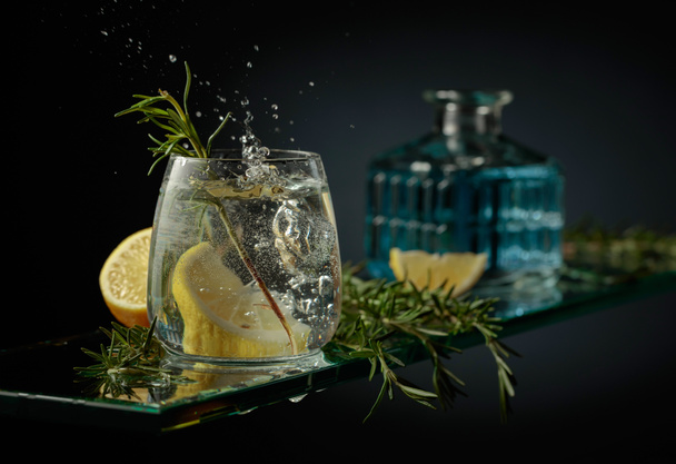 Gin tonic cocktail with lemon and rosemary. Slice of lemon fall in glass with a cocktail. - Photo, Image