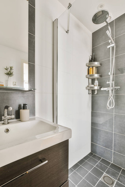 シャワーヘッドにグレーのタイルと白い器具を備えたモダンなバスルームがシンクの上の壁に取り付けられています - 写真・画像