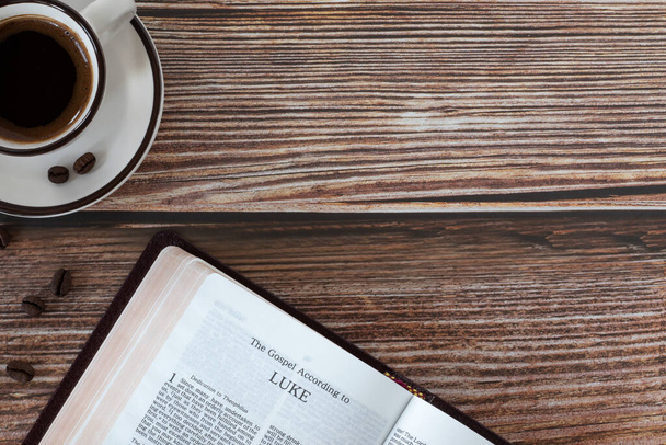Otwórz świętą księgę Ewangelia Łukasza z filiżanką kawy na drewnianym tle. Odbiór. Widok z góry stołu. Studiowanie Pisma Świętego, koncepcja biblijna. - Zdjęcie, obraz