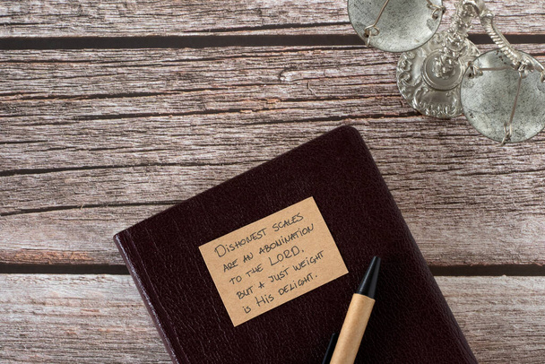 Biblical verse about honesty written on paper on top of holy bible book and ancient balance scales on wooden background. Top table view. - Photo, Image