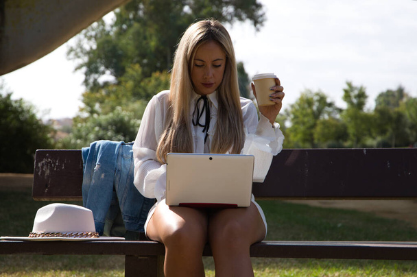 Portrét krásné mladé blond moderní oblečené ženy sedící na lavičce v parku s notebookem. Žena drží ekologický papírový šálek kávy nebo infuze. - Fotografie, Obrázek