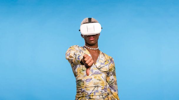 Black transgender person in stylish dress with VR helmet gesturing thumb down while exploring cyberspace against blue background - Photo, Image