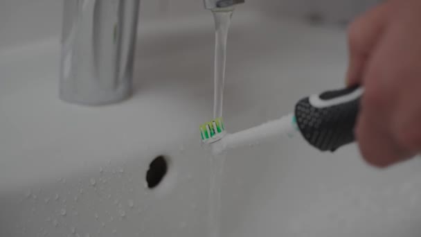 Mans Hände halten nach dem Zähneputzen eine elektrische Zahnbürste unter dem Wasserstrahl in der Spüle. Männchen wäscht die sonic power Zahnbürste mit Leitungswasser in der Badewanne. Mundhygiene am Morgen.  - Filmmaterial, Video