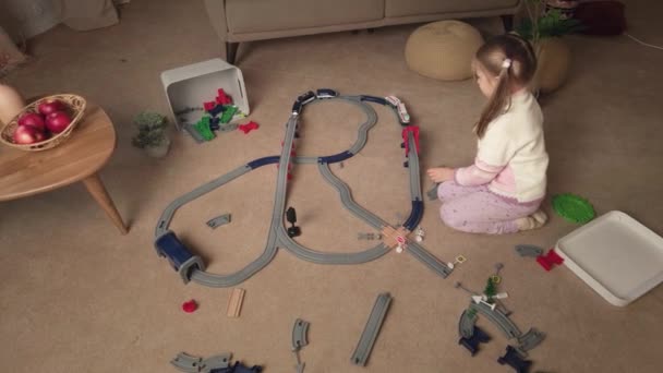 Feliz niña construye un ferrocarril de plástico de juguete y juega con un tren en casa. - Metraje, vídeo