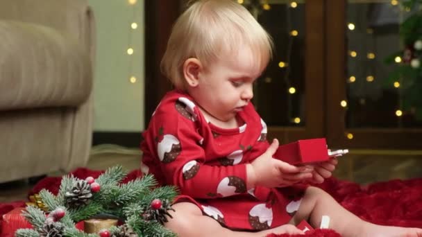 Mooie kleine een-jarige meisje met blond haar spelen kerstspeelgoed, bokeh bloemenslingers, kinderen en oudejaarsavond. - Video
