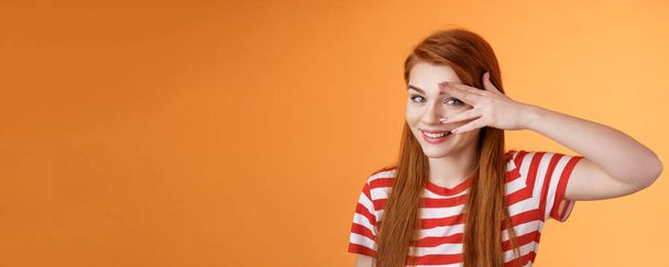 Charismatic lovely redhead lucky female, smiling cute, peek camera tender coquettish gaze, hold palm face, look through fingers, stand orange background optimistic, cheerful mood. Copy space - Photo, Image