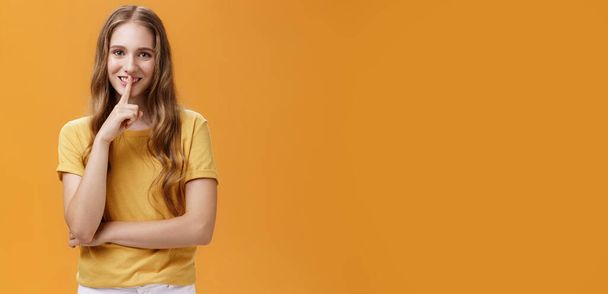 Girl wants keep secret betweet us. Portrait of charming good-looking slim pretty and young woman with long wavy hair and tattoo showing shh gesture with index finger over mouth against orange wall. - Photo, Image