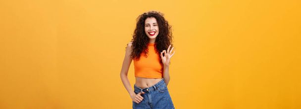 Charming young charistmatic woman. with curly hairstyle in cropped top and shorts showing okay or approval gesture smiling broadly liking awesome idea of friend where hang out over orange wall. - Photo, Image