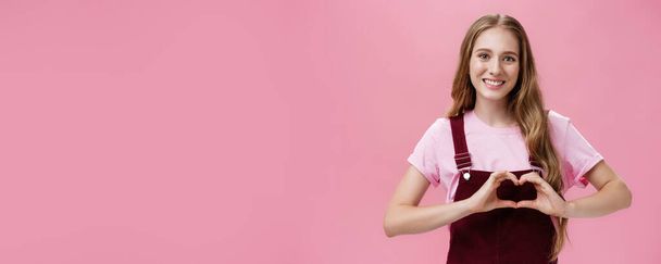 Girl loves family. Kind charming young woman in overalls with small tattoo on arm showing heart gesture over body and smiling lovely at camera expressing tender and cute attitude over pink wall. - Photo, Image