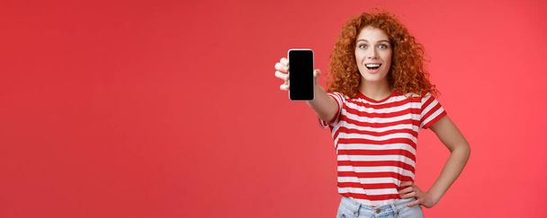 Fröhlich attraktive charismatische europäische rothaarige Mädchen lockige Frisur zeigen Smartphone-Bildschirm lächelnd glücklich fördern App Beratung empfehlen gute Anwendung Social Media Seite roter Hintergrund. - Foto, Bild