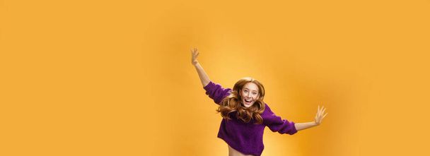 Expressive and enthusiastic carefree friendly joyful redhead woman in purple sweater and pants jumping joyfully with raised hands smiling yelling from happiness and joy against orange background. Copy - Photo, Image