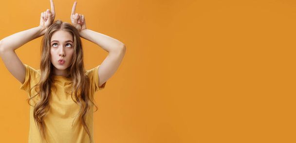 Lifestyle. GIrl going crazy from boredom making freaky funny faces fooling around squinting holding index fingers on head like horns sticking out tongue and folding lips aping over orange background. - Photo, Image