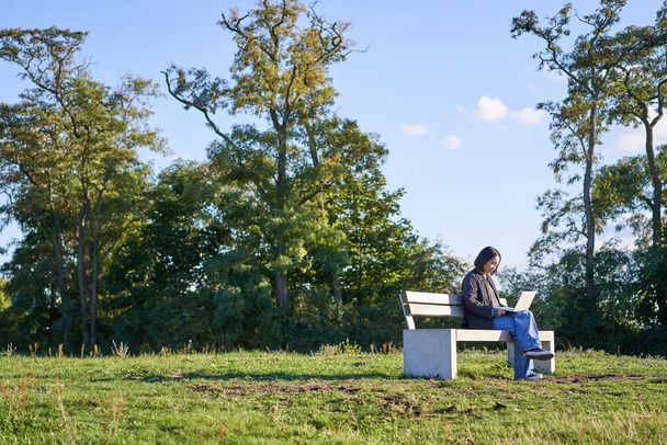 Vista lateral de una mujer joven sentada sola en el parque en el banco, usando su computadora portátil para estudiar o trabajar de forma remota desde el aire libre. - Foto, imagen