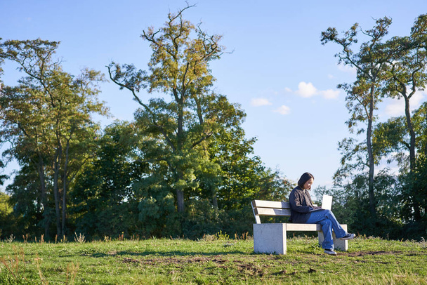 Πλευρική άποψη της νεαρής γυναίκας που κάθεται μόνη της στο πάρκο στον πάγκο, χρησιμοποιώντας το laptop της για να μελετήσει ή να εργαστεί εξ αποστάσεως από εξωτερικούς χώρους. - Φωτογραφία, εικόνα