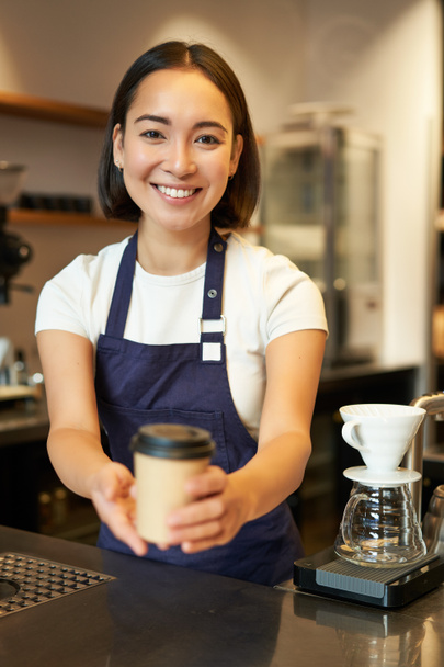 Улыбающаяся азиатка-бариста отдаёт приказы клиенту, держит чашку кофе на вынос, носит фартук, работает за прилавком в кафе. - Фото, изображение
