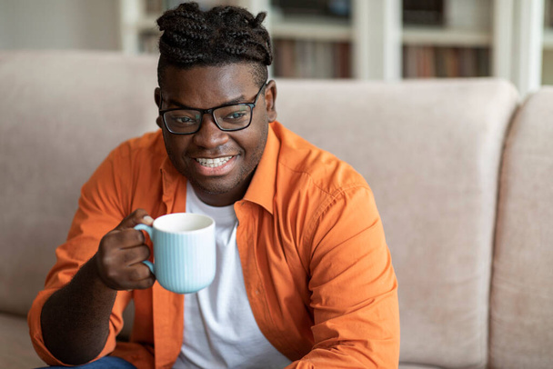 Uśmiechnięty przystojny pulchny czarny facet w luźnym stroju siedzi na kanapie z kubkiem kawy, pozytywny afrykański facet pijący herbatę w przytulnym salonie w domu, patrząc na przestrzeń na reklamę - Zdjęcie, obraz
