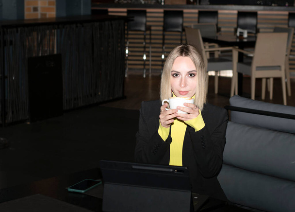 Mensen. Een prachtig blank blond meisje in een zwart jasje zit in een café met een kop koffie en communiceert vrolijk en positief online op een tablet. Concept. - Foto, afbeelding
