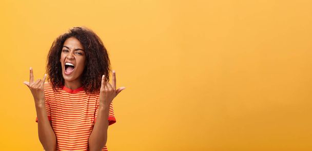 Cintura-para arriba tiro de asombrado feliz mujer afro-americana con estilo sintiendo impresionante mecedora en la fiesta gritando de alegría y satisfacción mostrando rock n roll gesto posando sobre fondo naranja. Copiar espacio - Foto, imagen