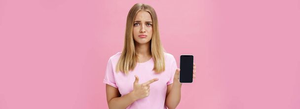 Θλιμμένη και λυπημένη νεαρή κοπέλα που δείχνει αρνητική ανατροφοδότηση για την εργασία της στο διαδίκτυο δείχνοντας την οθόνη του smartphone να στέκεται ανήσυχη και αναστατωμένη σε ροζ φόντο. Τεχνολογία και - Φωτογραφία, εικόνα