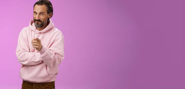 Homem barbudo adulto carismático bonito confiante com capuz rosa elegante convidando você a se juntar à empresa sorrindo auto-confiante indicando a escolha tomar a decisão de nos escolher, fundo roxo. - Foto, Imagem