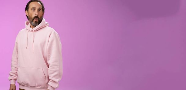 Beeindruckt sprachlos schockiert erwachsener bärtiger Mann graues Haar in coolem rosa Kapuzenpulli Faltlippen wow knallende Augen Kamera überrascht erstaunt fasziniert, stehend neugierig erstaunt lila Hintergrund. - Foto, Bild