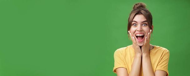 Portrait of happy delighted and surprised young feminine girl in yellow t-shirt pressing hands to cheeks from amazement and joy smiling broadly reacting to astonishing news over green background - Photo, Image