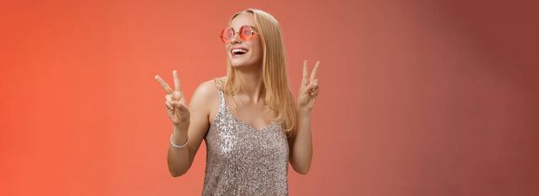 Positive charming european carefree stylish girlfriend having fun dance-floor show victory peace signs look up happily smile in sunglasses glamour glittering dress enjoy celebration, red background. - Photo, Image