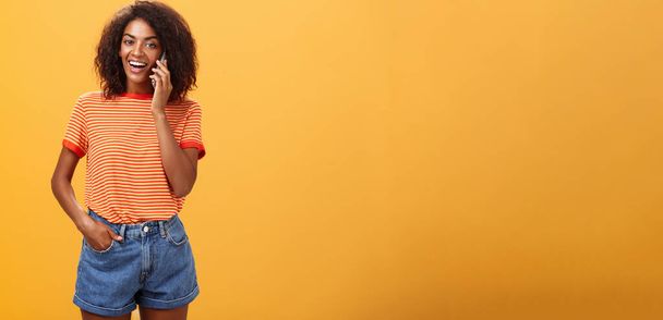 Indoor shot van stijlvolle zelfverzekerde vrouwelijke collega in trendy t-shirt en spijkerbroek houden smartphone in de buurt oor bellen vriend praten via mobiele telefoon terloops over meisjes spullen over oranje achtergrond - Foto, afbeelding