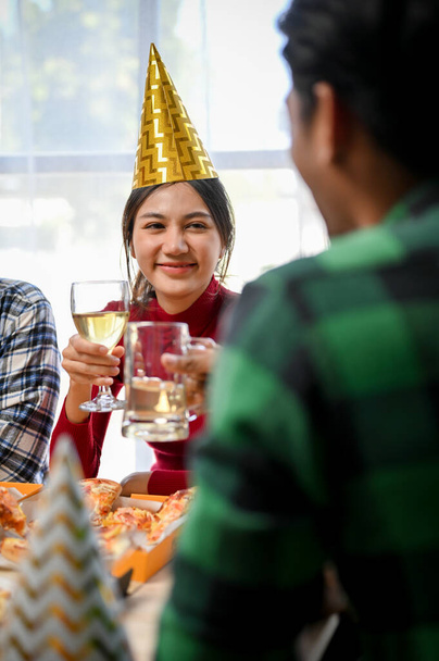 Portret, mooie jonge Aziatische vrouw met feestmuts, met een glas wijn, glimlachend en kijkend naar haar mannelijke vriend die op het feest aan het praten is. - Foto, afbeelding