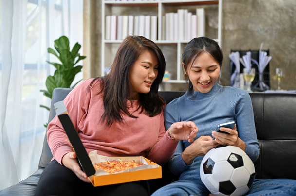 Twee charmante jonge Aziatische vriendinnen zitten op de bank in de woonkamer, het bekijken van een voetbalwedstrijd op een smartphone, het hebben van leuke tijd en pizza party thuis. - Foto, afbeelding