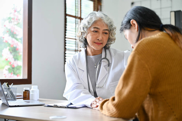 Profesional y cariñosa Asiática anciana médico hablando, teniendo una conversación seria con su joven paciente en el consultorio del hospital. - Foto, Imagen