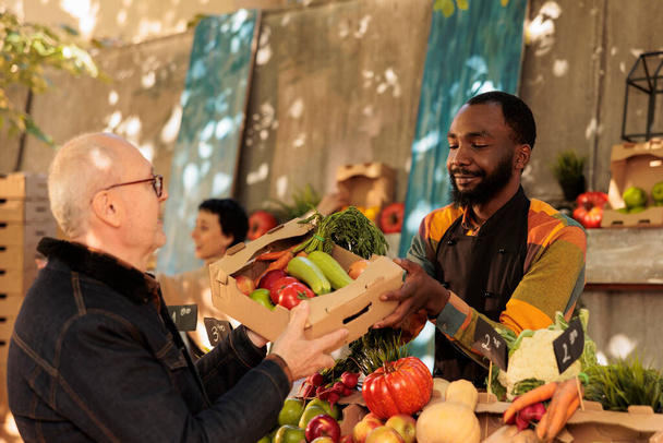 Старший покупает различные свежие сезонные фрукты и овощи на фермерском рынке осенью. Приятный улыбающийся афроамериканец владелец малого бизнеса, продающий свои домашние выращенные органические продукты - Фото, изображение