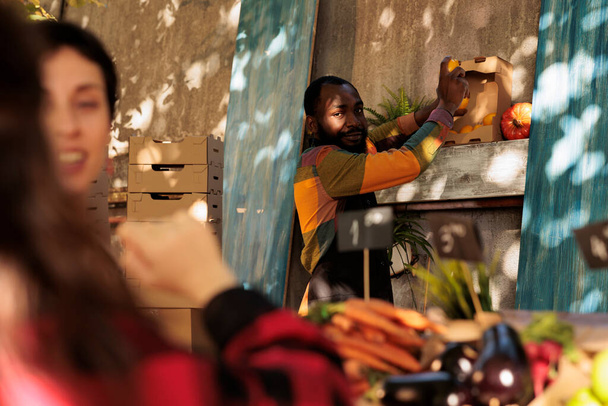 Afrikkalainen amerikkalainen viljelijät markkinoilla myyjä kuljettaa sitruunoita käsissä ja hymyillen asiakkaille, seisoo takana hedelmiä ja vihanneksia seistä. Ihmiset ostavat tuoretta bioluomua paikallisesti kasvatettuja tuotteita. - Valokuva, kuva