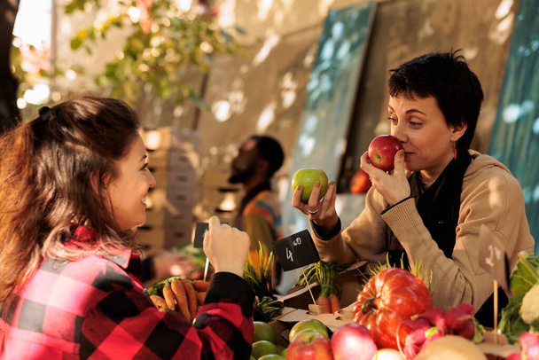 Farm to table. Female farmers market vendor smelling red apple while selling fresh organic fruits and vegetables to customer on sunny autumn day. Young happy woman buying locally grown healthy food - Photo, Image
