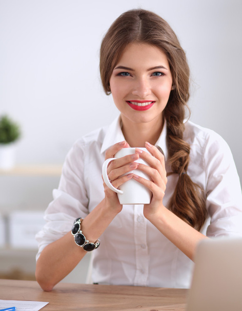 Young businesswoman sitting on the desk with cup in office - 写真・画像