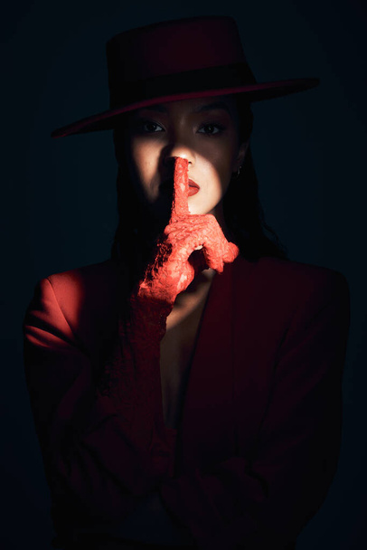 Fashion, silence and woman in red suit on a dark studio background. Quiet, spotlight and face portrait of beautiful, vintage or 90s female model in makeup cosmetics with shush hand gesture for peace - Photo, image