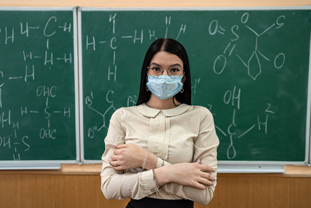 joven maestra en máscara protectora de pie cerca de pizarra. fórmulas químicas escritas en el aula - Foto, Imagen