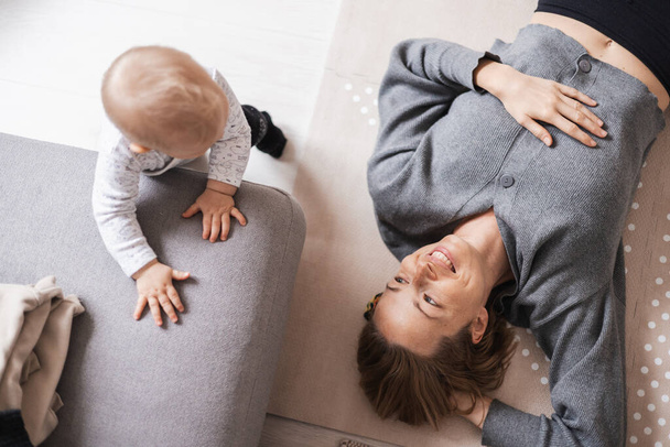 Felices momentos familiares. Madre acostada cómodamente en la alfombra de los niños jugando con su bebé viendo y suponiendo sus primeros pasos. Emociones humanas positivas, sentimientos, alegría - Foto, imagen