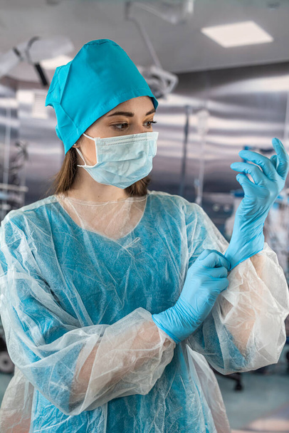 médico o enfermero con un abrigo azul se pone guantes de goma azul se prepara para examinar a un paciente en el quirófano. medicina. médicos y cirujanos de quirófano. - Foto, imagen