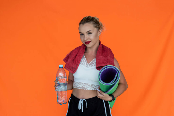 Μια νεαρή γυναίκα με ένα μπουκάλι αθλητικού νερού και ένα στρώμα γυμναστικής στέκεται σε ένα έγχρωμο φόντο. Αθλητική έννοια - Φωτογραφία, εικόνα
