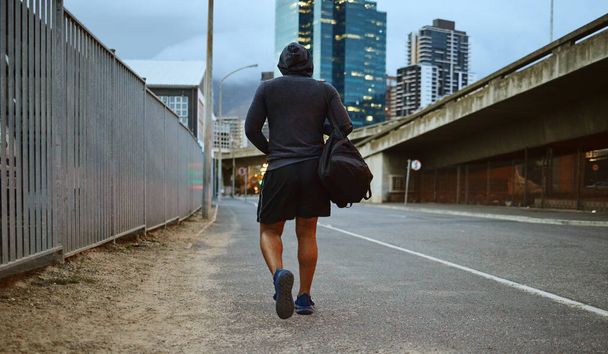 Fitness, hombre negro y caminar por la calle de la ciudad después de correr, ejercicio y entrenamiento de gimnasio con una sudadera con capucha por la noche. Espalda de un atleta masculino en Miami urbano para un paseo y entrenamiento cardiovascular con una bolsa de lona. - Foto, Imagen