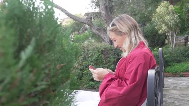 Una mujer lee las noticias en las redes sociales desde su teléfono en la primavera en el parque. - Imágenes, Vídeo